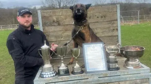 Humberside Police Quinta sits on a platform so is at the same level as her handler. There are five trophies and a certificate in front of her. She holds her head up high with her ears held back. PC Lusby holds her lead and stands on the left of the picture. He is dressed in black and wears a police cap.