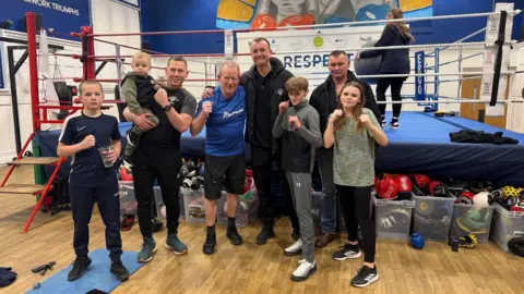 Mike Bromby A group of youngsters with four older men holding up their fists and standing next to a boxing ring. They are all wearing trainers and sporting wear.