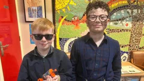 Seth and Danan. Seth has ginger hair, wearing sunglasses and a black bomber jacket, holding a toy squirter gun. Danan has brown curly hair, wearing glasses with the right glass cracked, a navy check shirt and navy suspenders.