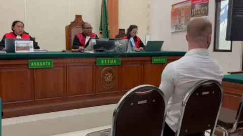 A picture of the Bali court room. The picture is taken behind Mr Parker who is sitting in front of three judges. The judges are wearing black shirts and red jackets with no sleeves. They are sitting at a large wooden desk.