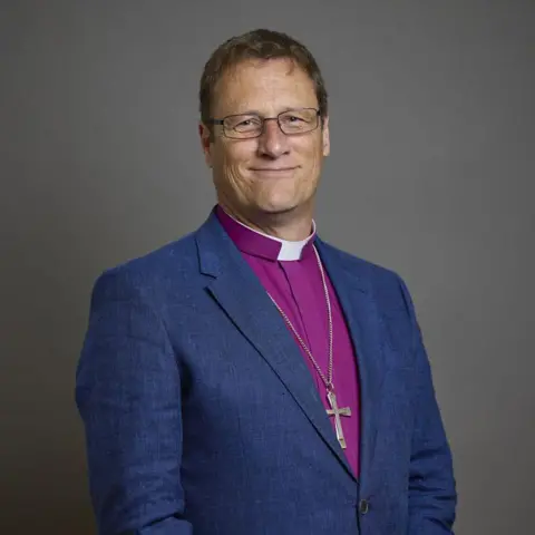House of Lords/Roger Harris A portrait of Bishop of Leicester, Martyn Snow, wearing a navy blue blazer with a pink shirt. He is smiling and wearing glasses, with a grey background.