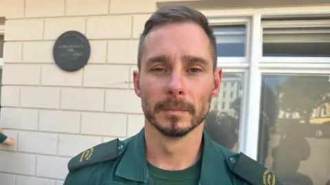 Tom Le Sauteur standing in front of a cream brick wall and window, wearing ambulance uniform.