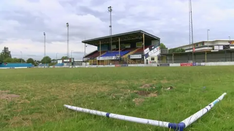 The Camrose. There is a large red and blue grandstand in front of a grass pitch.