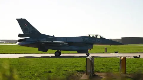 A F16 fighter aircraft in grey livery taxis along the runway at RAF Waddington