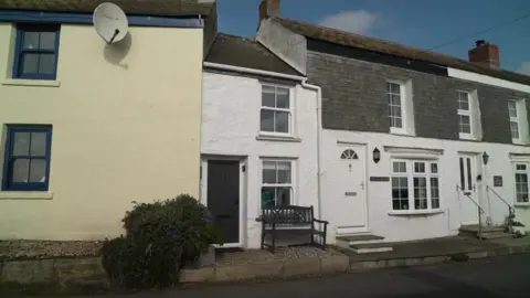 BBC The Doll's House sits in between a yellow and a second white house. The small white house has a grey bench outside it and a grey front door. 