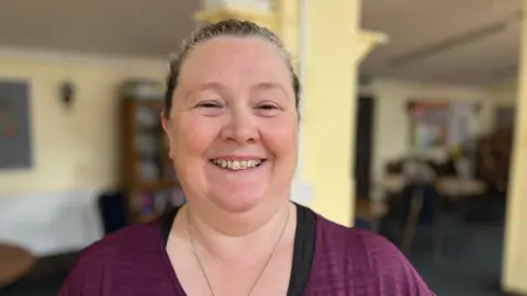 Aimee Dexter/BBC Carys Dale is standing in front of a wall. She has her hair in a ponytail and is wearing a purple and black T-shirt and a silver necklace.