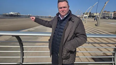 LDRS Councillor Gerard Walsh, wearing a brown jacket over a blue gillet and white shirt, points to the beach in Blackpool with the pier in the background across a large expanse of sea.