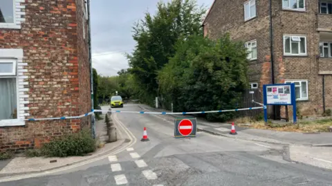 Jack Hadaway-Weller/BBC A street with what appears to be residential buildings on either side. There is blue and white police tape cordoning off access to the area and a red circle stop sign placed in the road. 