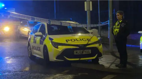 A female police officer stands next to a police car next to police tape connected between two lampposts, with other emergency vehicles seen in the background. 