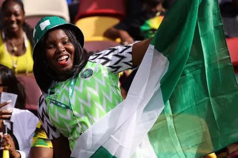 JACQUES FEENEY / OFFSIDE / GETTY IMAGES A women dressed in the Nigeria kit waves the Nigerian flag from the stands in the stadium.