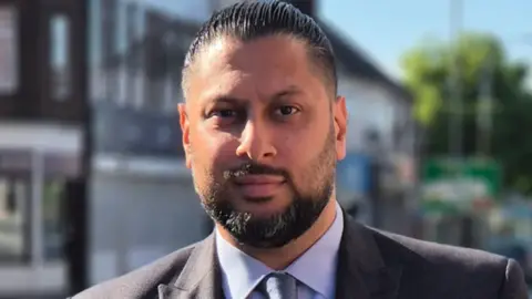 LDRS A man facing the camera. He has black hair and a black beard and moustache. He is wearing a grey suit with a blue shirt and tie.