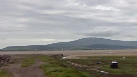 A Google Streetview showing the tide out at Duddon Estuary. There is a boat on a grassy area of dry land. Beyond that is a huge stretch of sand with a line of water - akin to a meandering stream - but the tide is a long way out of shot to the left. Beyond the wide estuary, in the background, hills gradually rise up from the left to a domed peak, creating what looks from distance like a rolling ridge. There are plenty of grey clouds but they are high and the day appears dry.