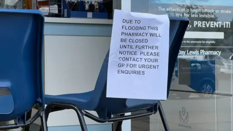 Stuart Woodward/BBC An A4 piece of paper attached to a blue plastic seat, which is positioned outside a GP surgery. The sign reads: "Due to flooding this pharmacy will be closed until further notice. Please contact your GP for urgent enquiries".