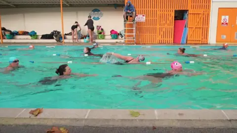 This image shows an outdoor swimming pool with several people swimming in marked lanes, while others are standing or preparing equipment beneath a covered area beside the pool.