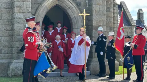 The choir, dressed in red and white robes, leaving the chapel while the trumpeters look on.