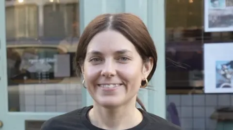 A woman with brown hair that is tied back smiles at the camera. She stands in front of a coffee shop wearing a black long sleeved t-shirt and gold hoop earrings. The shop behind her has a large front window and pale green painted door.