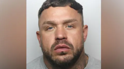 GMP Sebastian Jones has short brown hair and a beard. He wears a grey sweat shirt.