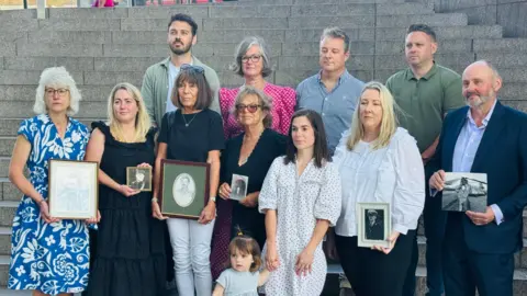 Chinook Justice Campaign Mrs Rawcliffe stands at the far left of a group of people who are also campaigning. They all look away from the camera. Some of them hold black and white photos of their relatives they lost in the crash. Some of the group stand up on steps above one other line of people. 