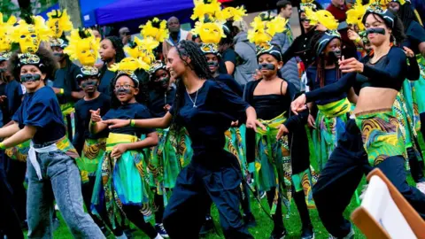 Stuart Whincup/BBC Schoolchildren dancing in costume at Middlesbrough's popular Orange Pip Market. They have yellow masks attached to their heads and their arms are outstretched. They are wearing jeans and blue trousers with green, and yellow skirts wrapped around their waists. They also have black stripes across their noses. 