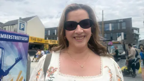Rachel Gilroy - a woman with long brown hair - is wearing a starfish necklace, a white frilly top with flowers on and is smiling in black sunglasses. Behind her you can see people gathered in a street and a bar and benches in the background.