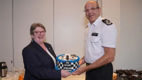 PC Karen Giles, standing on the left, is holding a cake by Assistant Commissioner Matt Twist, standing on the right. PC Karen Giles is wearing a dark navy blazer with a white blouse underneath and glasses. The Assistant Commissioner is wearing a white shirt with a police officer badge that is visible on the left shoulder, along with black trousers. The cake is decorated with a white and black checkered design and blue icing. In the background is a cream wall and a white table with cups, plates, drinks and party food. 