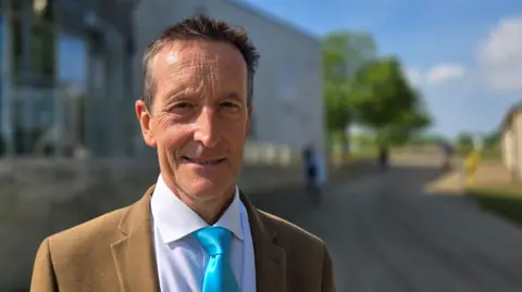 Paul Moseley/BBC Nick Taylor is wearing a light brown suit jacket on top of a white shirt and turquoise tie
