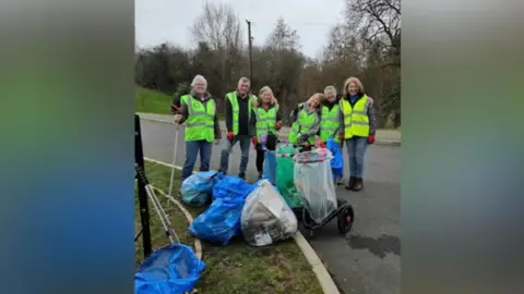 Carol Ludwig A group of male and female litter pickers stand by the side of a road with bags of rubbish in front of them. They all have florescent jackets on and are smiling at the camera.