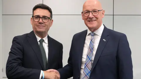 Andy Burnham, a man with thick dark hair and black-rimmed spectacles, is shaking hands with John Swinney, a man with a bald head and dark-rimmed spectacles. Burnham is in a dark blue suit with white shirt and solid green tie while Swinney is in a dark blue suit with white shirt and a blue, red and grey tartan tie. They are both smiling against a grey backdrop.