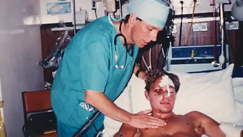 Headway Dominic Hurley laid in a hospital bed with a bandage on his face. There is a doctor wearing scrubs in a hospital ward.