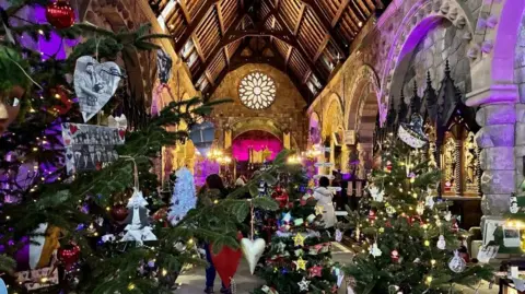 Ruth Walsh Un interior de iglesia decorado con múltiples árboles de Navidad adornados con adornos y luces, bajo un alto techo de madera con arcos de piedra y una vidriera circular en el extremo más alejado.