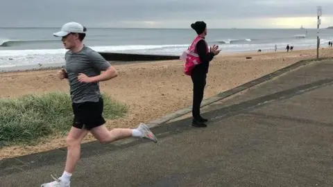 Charles McManus An image of Charles McManus running wearning a grey top and black shorts with the beach and the sea behind 