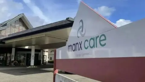 A closeup of a large white sign outside the entrance to Noble's Hospital, which has grey and green writing that reads Manx Care.