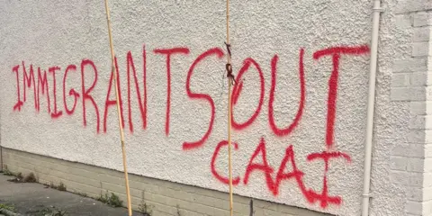 A white gable wall with the words: 'Immigrants out' sprawled in red paint.
A section of the message has been blurred.
A pink rose bush with some flowers can be seen at the side.