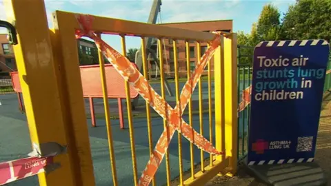 BBC Tape saying 'toxic air' is wrapped around a gate at a Birmingham park near a sign that says 'Toxic air stunts lung growth in children'