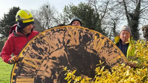 Reverend Daniel Whiffin Three workmen standing behind a large clock face, which is visibly worn and old. Trees are behind them and a bush is in front of them.
