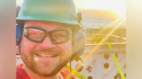 Phil Perry Offshore worker Phil Perry in hard hat, smiling at camera on an offshore platform, with sea and sunshine behind him.