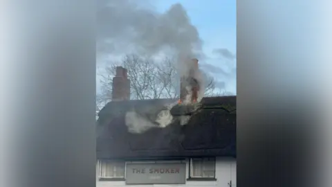 Shaun Gee A thatched roof with two chimneys and a pub sign which says the smoker on the front of the white building, between two windows, fire is coming from one of the chimneys