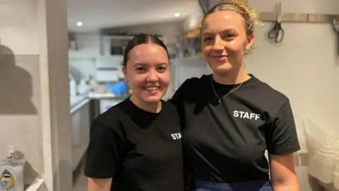 Amber e Bella estão usando camisetas pretas com "Funcionários" neles. Um tem cabelos castanhos amarrados para trás, o outro tem cabelos loiros encaracolados amarrados para trás. Eles estão de pé lado a lado em uma cozinha.