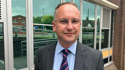 A man wearing a grey suit with a blue shirt and a red and blue tie, stood behind a modern glass-fronted building.
