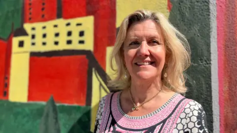 Sue Dougan/BBC Head and shoulders image of Julie Kerr standing in front of a colourful wall. Julie has shoulder length blonde hair and is wearing a necklace and a pink, white and black patterned dress.