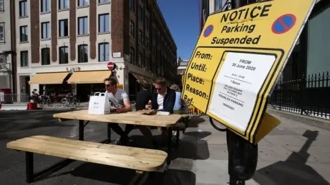 PA Media People eat lunch at tables and benches placed in the roadway adjacent to a suspended parking bay in Golden Square, Soho