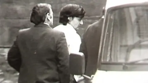 A black and white still from TV footage showing Peter Sullivan, who has thick black hair and is wearing a white shirt, being led to a police van in handcuffs.