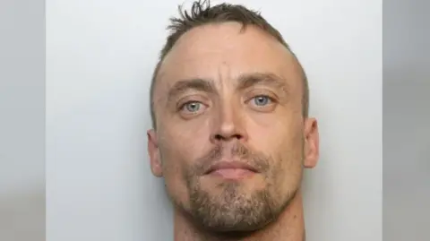 Avon and Somerset Police Custody picture of Ashley Billing. He is staring directly into the camera, and has short light brown hair and stubble. He is standing against a plain grey background and a neck tattoo is faintly visible on the right side of his neck. 