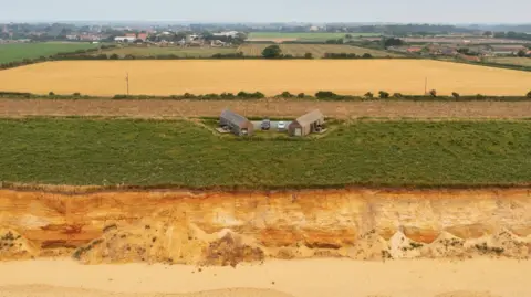 Anne Jones An aerial shot showing eroding sandy cliffs in the foreground, then green land on the cliff and a small clutch of buildings