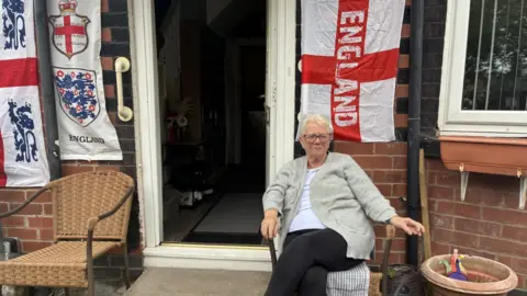 Mary, a woman with short white hair and glasses, sits on a chair in front of a house covered with various England flags.