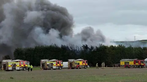 Nottinghamshire Fire and Rescue Service Smoke rising behind trees with fire engines in attendance at a site in Gunthorpe, Nottinghamshire.