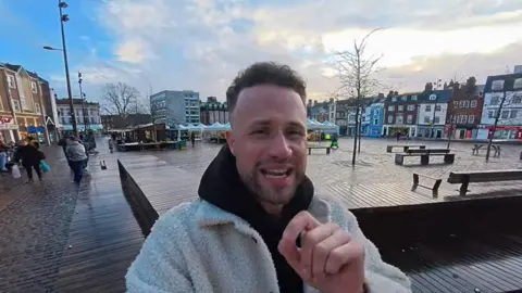 Jamie Benkert Jamie Benkert speaking to his camera in Northampton market square. He has black hair and stubble and is wearing a white fleecy coat. There are shoppers behind him.