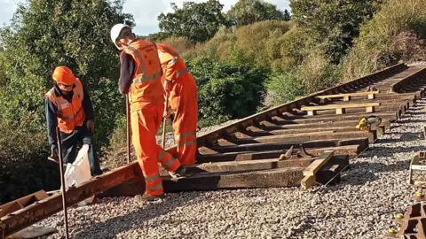 North Dorset Railway Three men in orange hi-viz overalls and hard hats manoeuvring a section of steel railway track into position. Behind them a new section of track and sleepers are already in place.