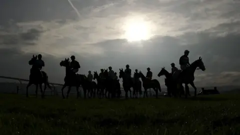 Getty Images A group of jockeys on their horses are pictured near the start line at Taunton Racecourse. They are silhouetted dark against the sunny sky with light clouds also visible