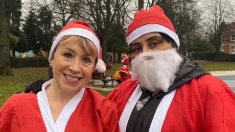 Two women are looking directly towards the camera. They are in a green park dressed as santa. They both have red and white outfits on with the person on the left with no beard and the person on the right with on.